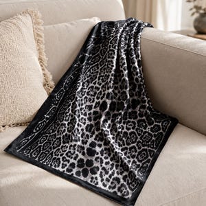 May include: A black and white leopard print throw blanket draped over a beige sofa. The blanket features a repeating pattern of dark spots on a light background, with a black border. A beige fringed pillow is visible.