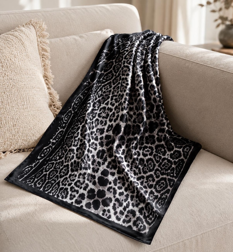 May include: A black and white leopard print throw blanket draped over a beige sofa. The blanket features a repeating pattern of dark spots on a light background, with a black border. A beige fringed pillow is visible.