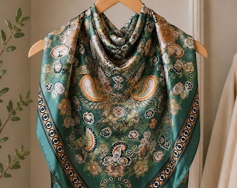 May include: A square silk scarf with a paisley pattern in shades of green, gold, and cream. The scarf is displayed on a wooden hanger. The border features a repeating geometric design in a darker green.