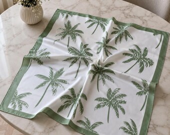 May include: White square scarf with a green palm tree pattern and a green border. The scarf is made of a soft, silky material and is displayed on a marble table.
