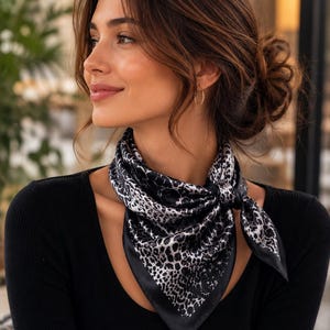 May include: A black and white leopard print silk scarf tied around the neck. The scarf has a black border and is knotted in the front. The person is wearing a black long-sleeve shirt.