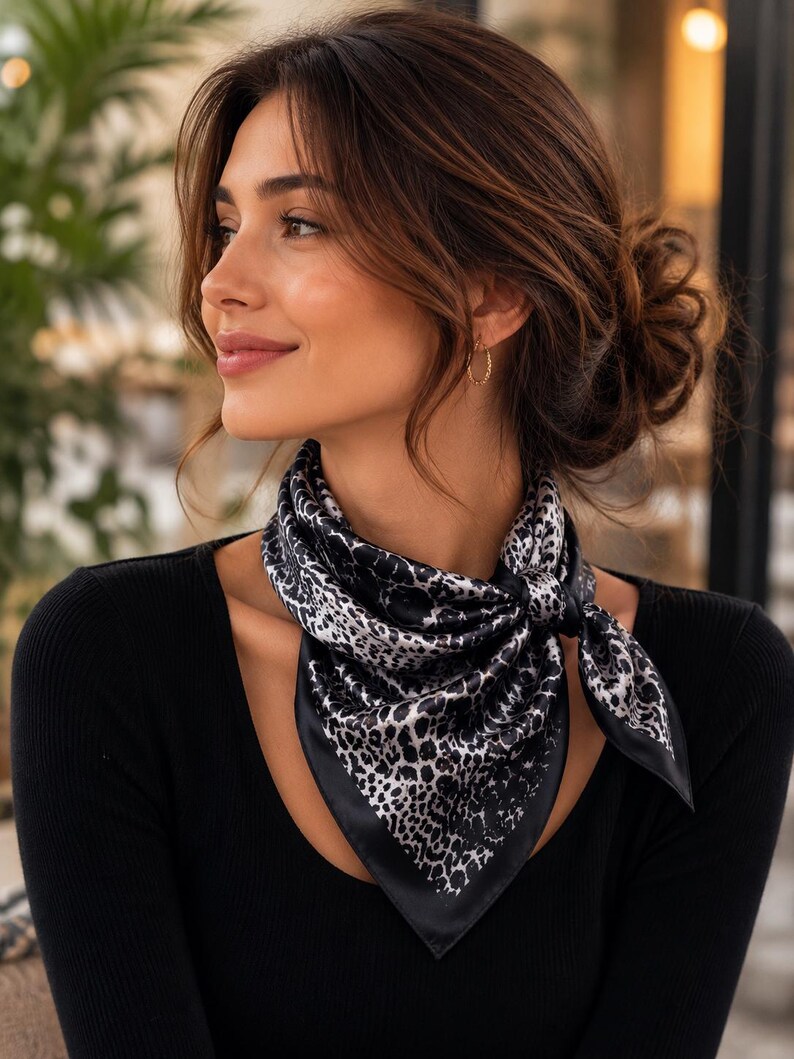 May include: A black and white leopard print silk scarf tied around the neck. The scarf has a black border and is knotted in the front. The person is wearing a black long-sleeve shirt.