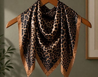 May include: A square silk scarf with a leopard print design. The scarf features a black and tan animal print pattern with a solid tan border. The scarf is displayed on a wooden hanger against a neutral background.