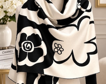 May include: A black and white scarf featuring a large-scale floral pattern with bold, stylized flower shapes. The scarf is draped over a mannequin, displaying a high-contrast design with solid black petals and outlines on a cream-colored background.