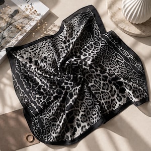 May include: A black and white leopard print silk scarf. The scarf is folded, revealing the pattern and a dark border. It rests on a neutral surface, alongside a book and decorative items.