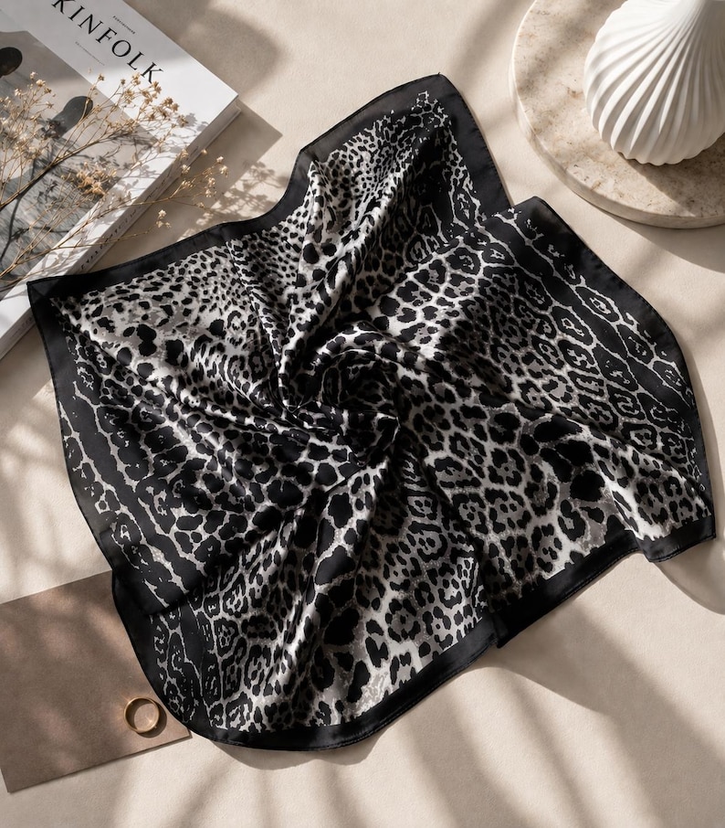 May include: A black and white silk scarf with a leopard print design. The scarf has a dark border and is folded, showcasing the pattern. The scarf is on a neutral surface with a book and decorative items.