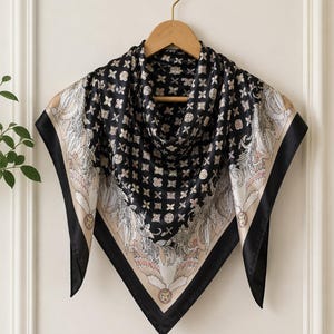 May include: A square black silk scarf with a geometric pattern and decorative border. The scarf features a repeating pattern of small shapes and a detailed design on the corners with cream, pink, and black accents. It is hanging on a wooden hanger.