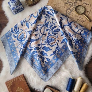 Op de afbeelding: Een vierkante zijden sjaal met een witte achtergrond en een blauw en bruin bloemenpatroon. De sjaal heeft een blauwe rand met een geometrisch ontwerp. De sjaal wordt gepresenteerd op een witte imitatiebont oppervlak.