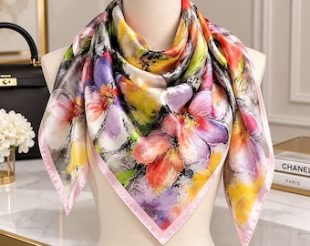May include: A square silk scarf featuring a vibrant watercolor-style floral print in shades of pink, yellow, purple, and green. The scarf has a solid light pink border and is draped over a mannequin, displaying a smooth, glossy finish.