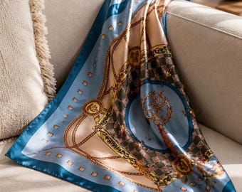 May include: A silk scarf with a blue border and a beige and blue center. The scarf features a chain and key design with gold accents. The scarf is draped over the arm of a beige sofa.
