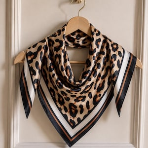 May include: A square scarf featuring a tan and black leopard print pattern. The scarf has a wide border consisting of a white stripe flanked by thin brown lines and a solid black edge. The fabric has a smooth, satin-like sheen and is draped.