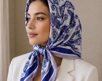 May include: A blue and white paisley patterned headscarf tied at the neck. The scarf has a blue border and is worn over the head. The scarf is paired with a white blazer and top.