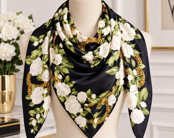 May include: A square scarf featuring a black background with a pattern of white roses, green leaves, and gold chain links. The scarf is draped around a mannequin neck, displaying a floral and metallic design with a smooth, satin-like finish.