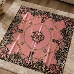 May include: A square pink scarf with a gold chain and black ornate design. The border features a leopard print pattern. The scarf is made of a shiny, silky material.