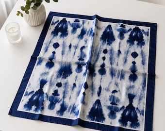 May include: A square, white and blue tie-dye patterned scarf with a dark blue border. The scarf is folded on a white surface. A small white vase with greenery and a candle in a glass holder are in the background.