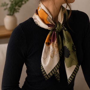 May include: A silk scarf with a floral pattern in shades of cream, orange, green, and burgundy is knotted around the neck. The scarf has a striped border and is worn over a black long-sleeved top. The scarf adds a touch of elegance.