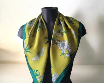 May include: A square, yellow scarf with a green border, adorned with white floral and butterfly patterns. The scarf is draped over a black mannequin, showcasing its design and color contrast. The scarf's design includes white flowers and butterflies.
