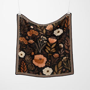 May include: A square scarf with a floral design. The scarf has a black base, adorned with peach, brown, and white flowers and leaves. It has a border with black and peach stripes. The scarf is hanging.