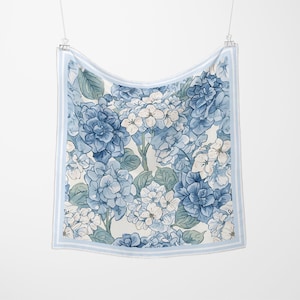 May include: Square scarf with a floral hydrangea pattern in shades of blue and white. The scarf has a light blue border with two stripes. The scarf is hanging from a metal clip.