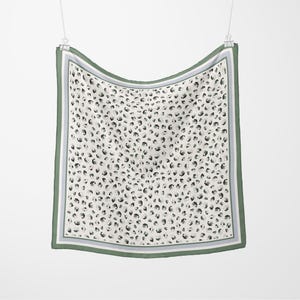 May include: A square scarf featuring a cream background with a black and grey animal print. The scarf has a green border with a grey and white stripe. It is suspended from a silver clip.