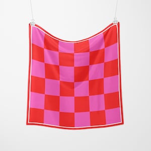 May include: A square scarf featuring a vibrant checkerboard design in red and pink. The scarf is edged with a thin white border and is hung from a metal clip. The fabric looks lightweight, suitable for different styling choices. The scarf measures approximately 70 cm x 70 cm.