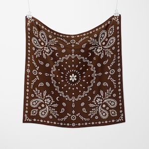 May include: A brown bandana with a white paisley and floral design. The square cloth features a central floral motif surrounded by a circular pattern of stars and crescent moons. The edges are adorned with a repeating floral border.