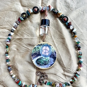 May include: A circular wooden plaque with a picture of a well with a green lid and a symbol above it. The plaque is hanging from a copper wire with a small glass bottle and a beaded necklace made of various colored stones.