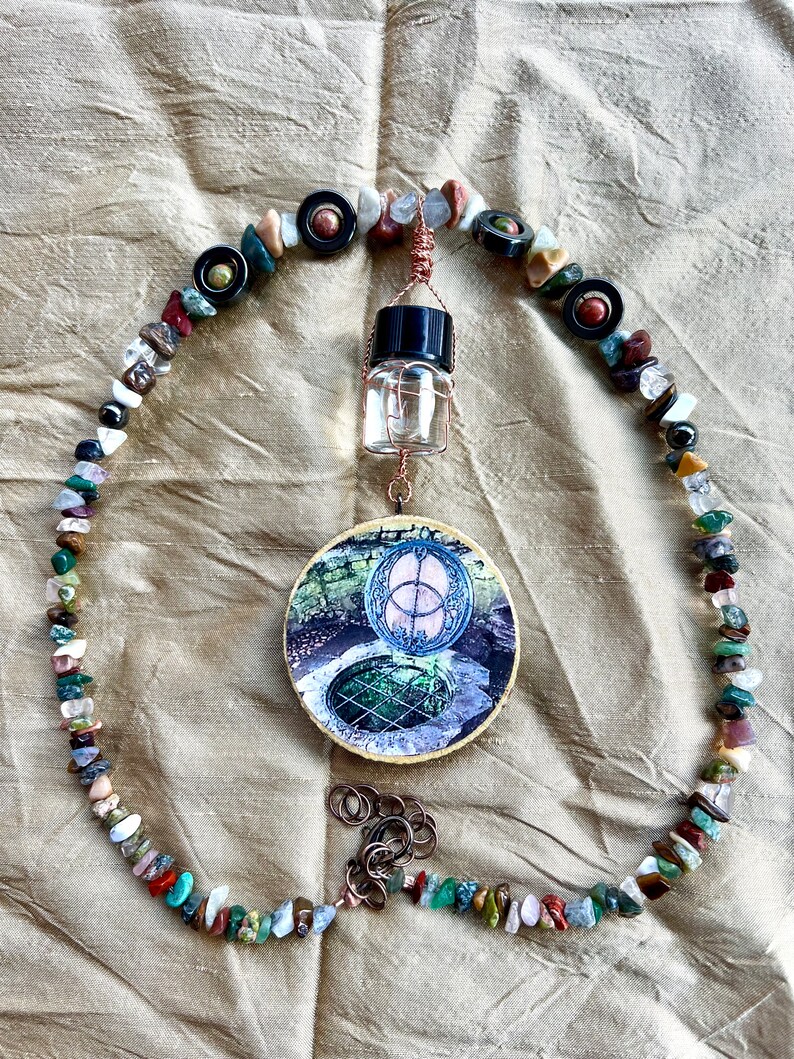 May include: A circular wooden plaque with a picture of a well with a green lid and a symbol above it. The plaque is hanging from a copper wire with a small glass bottle and a beaded necklace made of various colored stones.