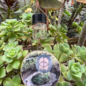 May include: A round wooden pendant with a picture of a well with a circular design on top. A small glass bottle with a black lid hangs from the pendant.