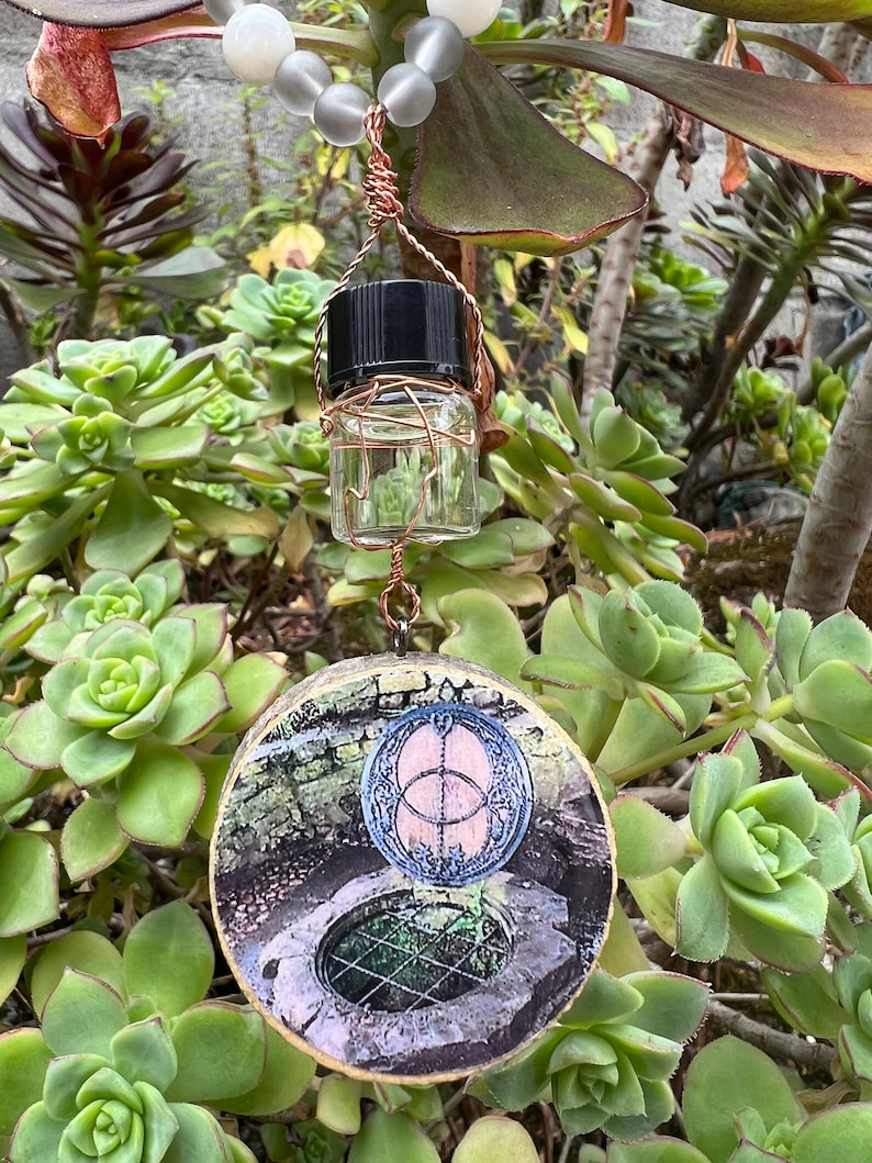 May include: A round wooden pendant with a picture of a well with a circular design on top. A small glass bottle with a black lid hangs from the pendant.