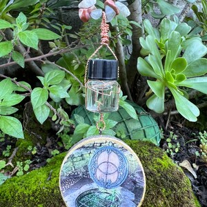 May include: A round wooden pendant with a picture of a well with a circular design in the center. The pendant is attached to a copper wire with a small glass bottle with a black lid.