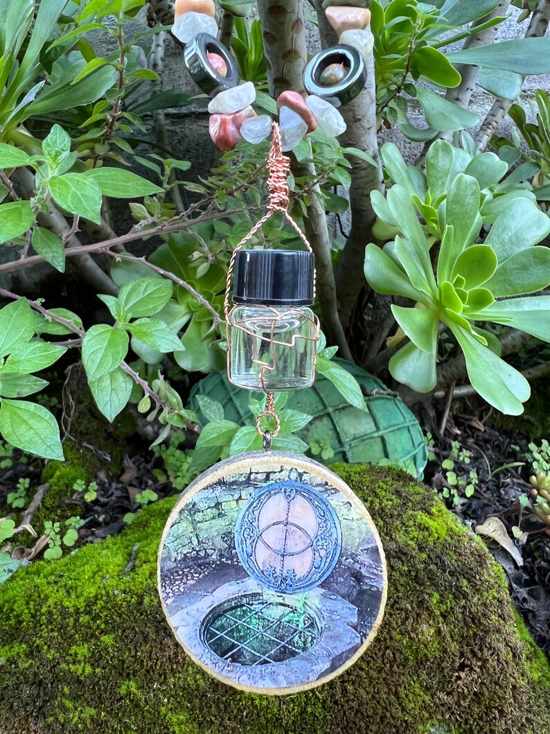 May include: A round wooden pendant with a picture of a well with a circular design in the center. The pendant is attached to a copper wire with a small glass bottle with a black lid.