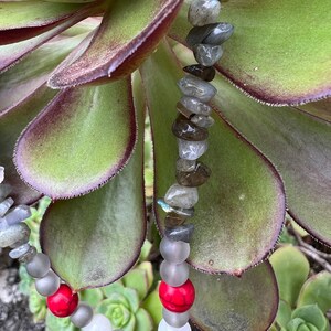 May include: A necklace made with grey and white beads, red beads, and grey and brown stones. The necklace is lying on a green succulent plant.