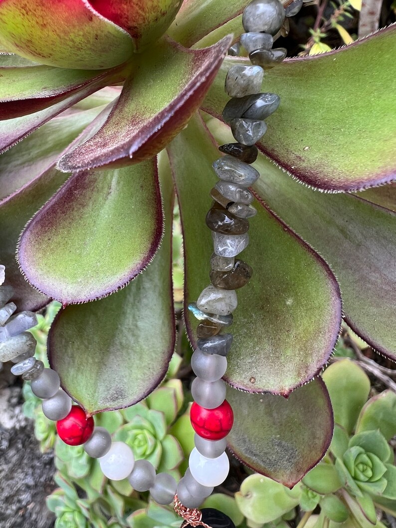 May include: A necklace made with grey and white beads, red beads, and grey and brown stones. The necklace is lying on a green succulent plant.