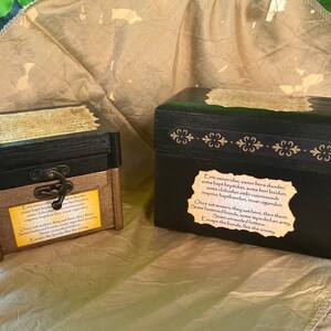 May include: Two wooden boxes with gold accents and a black top. The boxes have a decorative design on the front and a label with text that reads: "L'vis sazun idai, sazun hera duoder, suma hapt heptidus, suma heri lesidun, suma chubedun umbi coniund, princ hapthandun, iar vigandun. Once sat women, they sat here, then there. Some fastened bands, some impeded an army. Escape the bonds, flee the enemy."