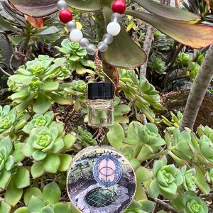 May include: A necklace with grey, white, and red beads, a small clear glass bottle with a black cap, and a round wooden pendant with a circular design and a picture of a well.