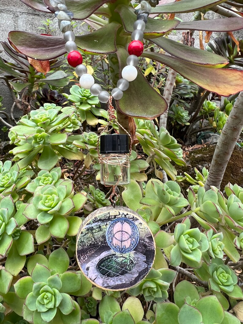 May include: A necklace with grey, white, and red beads, a small clear glass bottle with a black cap, and a round wooden pendant with a circular design and a picture of a well.