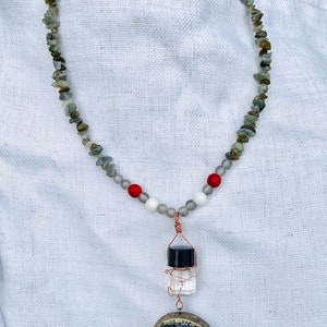 May include: A necklace made of grey and green stones with red and white beads. The necklace has a pendant with a small glass bottle and a round wooden disc with a picture of a well.