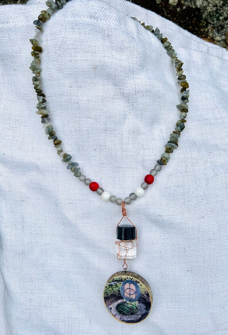 May include: A necklace made of grey and green stones with red and white beads. The necklace has a pendant with a small glass bottle and a round wooden disc with a picture of a well.