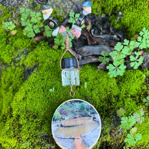 May include: A necklace made of various stones and beads, including green, red, and black, with a small glass bottle pendant and a round image of a stone fountain.