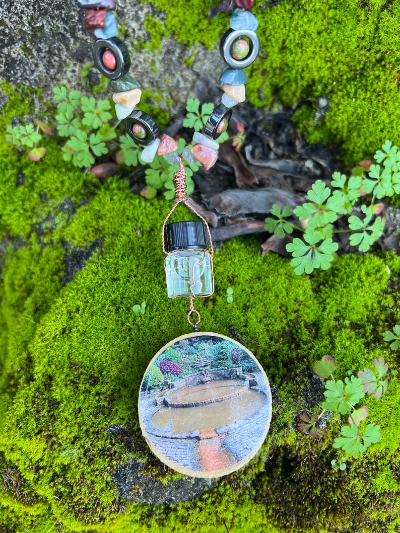 May include: A necklace made of various stones and beads, including green, red, and black, with a small glass bottle pendant and a round image of a stone fountain.