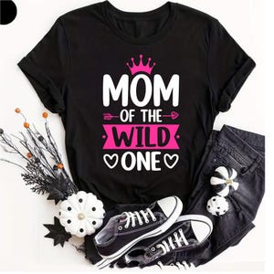May include: Black t-shirt featuring the text "MOM OF THE WILD ONE" in white and pink, with crown and arrow details. The shirt is styled with black jeans and black and white trainers. Decorative pumpkins and foliage are also present.