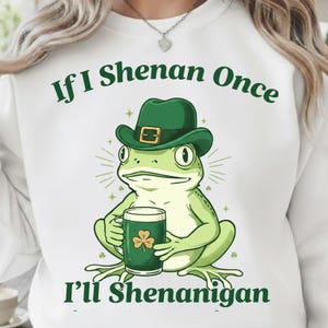 Pode incluir: Camisola branca com um sapo verde usando um chapéu verde e segurando uma caneca de cerveja com um trevo. O texto diz "If I Shenan Once I'll Shenanigan" em verde. O desenho está centrado na frente da camisola.