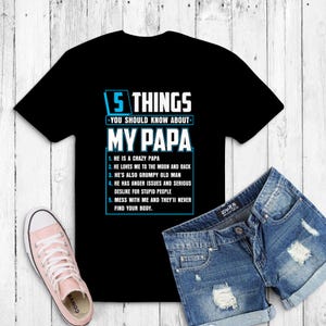 May include: Black t-shirt with the text "5 THINGS YOU SHOULD KNOW ABOUT MY PAPA" in white and blue. The shirt includes a list of humorous statements about a father. A pair of denim shorts and a pink sneaker are also in the image.