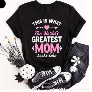 May include: Black t-shirt with the text "This Is What The World's Greatest Mom Looks Like" in white and pink. The shirt is paired with black jeans and black and white trainers. Decorative pumpkins and branches are also in the image.