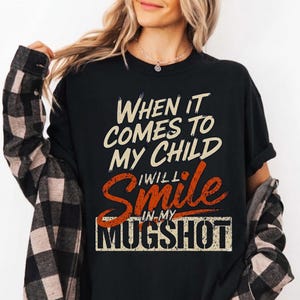 May include: Black t-shirt with the text "WHEN IT COMES TO MY CHILD I WILL SMILE IN MY MUGSHOT" in white and orange lettering. The shirt is worn by a person with long blonde hair.