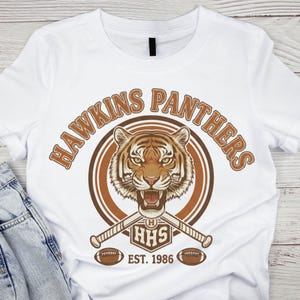 May include: White t-shirt featuring a tiger graphic with the words "HAWKINS PANTHERS" in brown arched text. Below the tiger are crossed baseball bats, the letters "HHS", and the text "EST. 1986". Two footballs are below the bats.