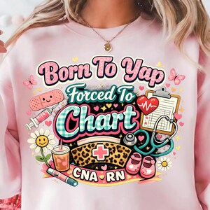 May include: Pink sweatshirt with a colorful graphic that reads "Born To Yap, Forced To Chart." The design includes medical-themed illustrations such as a bandage, syringe, stethoscope, and nursing cap. CNA and RN are also displayed.