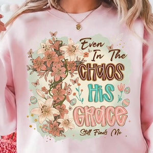 Könnte beinhalten: Hellrosa Sweatshirt mit floralem Kreuz-Design. Das Kreuz ist mit pfirsichfarbenen Blumen und grünen Ranken verziert. Der Text lautet "Even In The Chaos His Grace Still Finds Me" in verschiedenen Farben und Schriftarten.