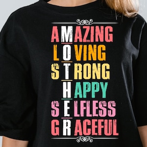 May include: Black t-shirt with the words "AMAZING, LOVING, STRONG, HAPPY, SELFLESS, GRACEFUL" in a colorful, stacked design. The text is in various colors, including pink, yellow, and teal. The shirt has a simple, classic crew neck.